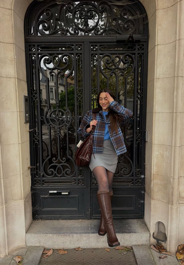 Une femme portant une veste à carreaux, une jupe grise et des bottes brunes franchit un portail en fer noir orné, avec un sac brun en bandoulière.