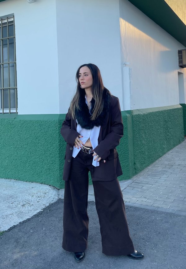 Blazer marrón de gran tamaño, collar de piel negra, blusa blanca y pantalones anchos negros. Los accesorios incluyen un cinturón plateado y joyas. Fondo de pared verde.