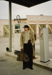 Beige trenchcoat över en stickad tröja, svarta vida byxor. Håller en mönstrad duffelväska och står nära bensinstationspumpar.