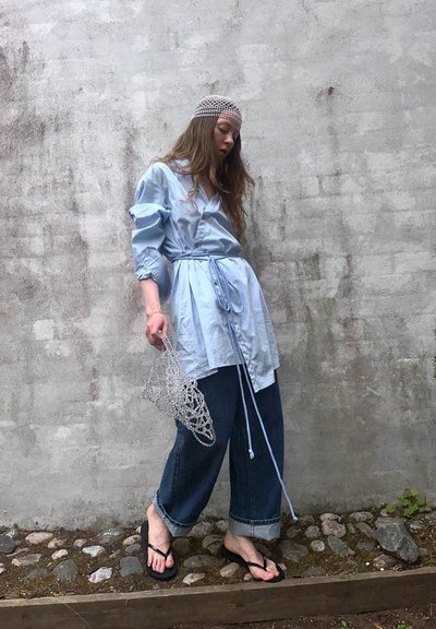 Camisa azul claro con botones y cinturón, combinada con pantalones vaqueros oscuros de pierna ancha. Sostenido un bolso transparente con patrón, usando chanclas negras.