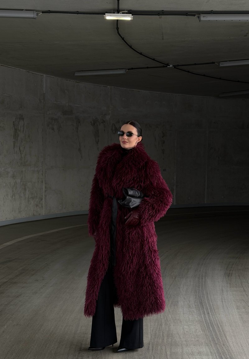 Maroon faux fur coat with a high collar, belted waist, and long sleeves. Black turtleneck, wide-leg black pants, and leather gloves.