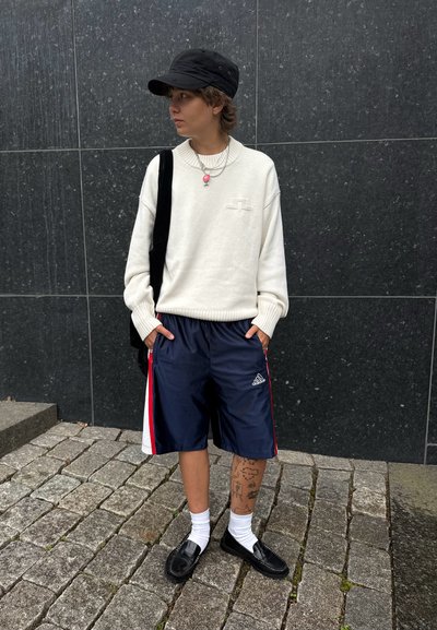 Suéter blanco, pantalones cortos navy con rayas rojas, gorra negra, zapatos negros, calcetines blancos, collares en capas y tatuajes visibles en una pierna.