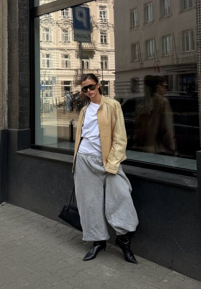 Mujer con chaqueta de cuero beige, camisa blanca, pantalones anchos grises, botas negras y gafas de sol, de pie en una acera de la ciudad junto a una ventana reflectante.