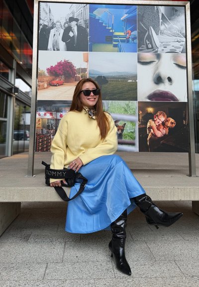 Suéter amarillo de punto, falda azul de satén, botas negras hasta la rodilla y un bolso negro con el texto "TOMMY". El fondo presenta un collage fotográfico.
