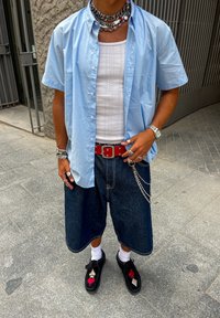 Persoană îmbrăcată cu o cămașă cu mânecă scurtă de culoare albastru deschis peste un maiou alb, pantaloni scurți din denim închis, pantofi negri cu simboluri de cărți de joc și bijuterii suprapuse, stând în aer liber.