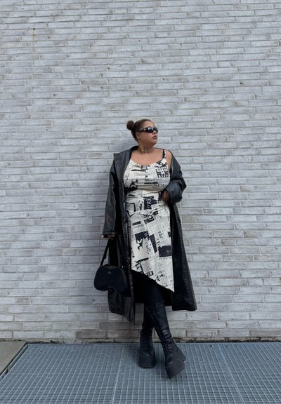 Abrigo trench negro brillante sobre un vestido crema con estampado de periódico, combinado con botas de plataforma negras hasta la rodilla y un pequeño bolso negro.