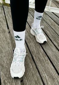 Baskets de sport blanches avec des accents vert clair, associées à des chaussettes blanches à côtes avec le texte "BERLIN" et un logo verts, portées avec un legging noir.