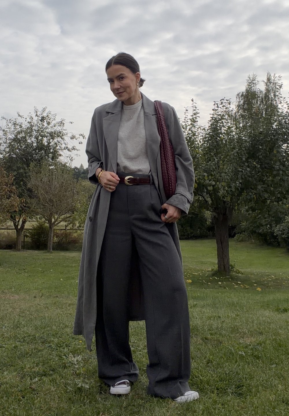 Szary płaszcz trench, jasnoszara sweter, szerokie szare spodnie, brązowy pasek i bordowa pleciona torebka. Białe sneakersy, tło z zielonej trawy.
