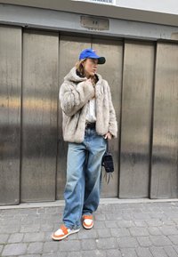 Kapuzenjacke aus Faux-Fur in hellbeige, im Oversized-Schnitt; kombiniert mit hellblauen, weit geschnittenen Jeans, weißer Sneakers mit orangefarbenen Akzenten und einer blauen cap.