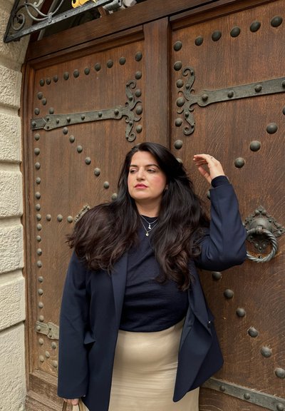 Mujer con cabello largo y oscuro y collares en capas se encuentra frente a una gran puerta de madera con clavos metálicos decorativos y un picaporte redondo de metal.