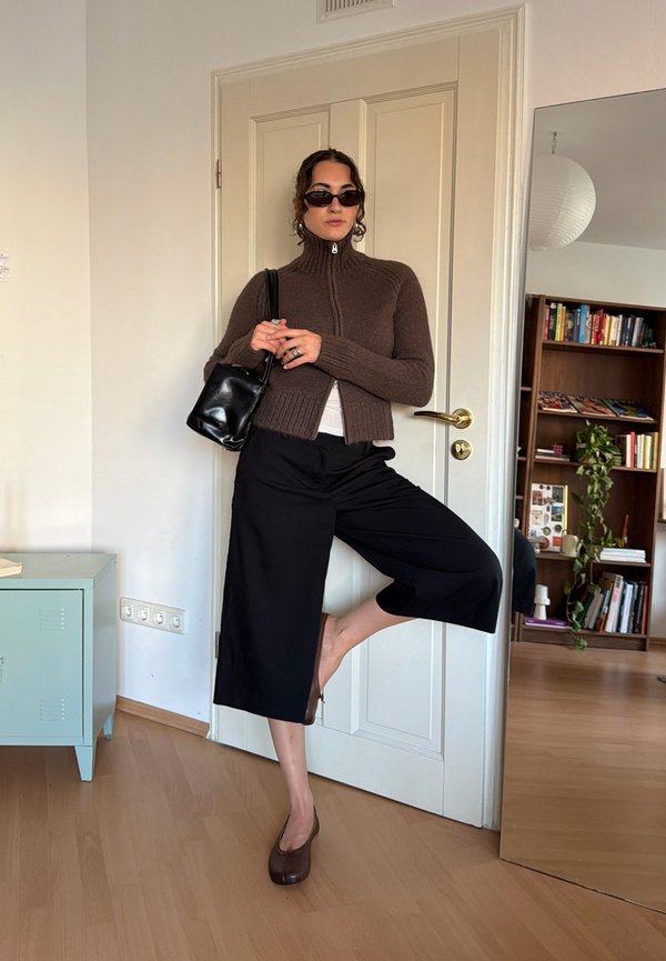 Mujer de pie sobre una pierna frente a una puerta cerrada, lleva gafas de sol oscuras, suéter marrón con cremallera, pantalones anchos negros y zapatos marrones, sosteniendo una bolsa negra.