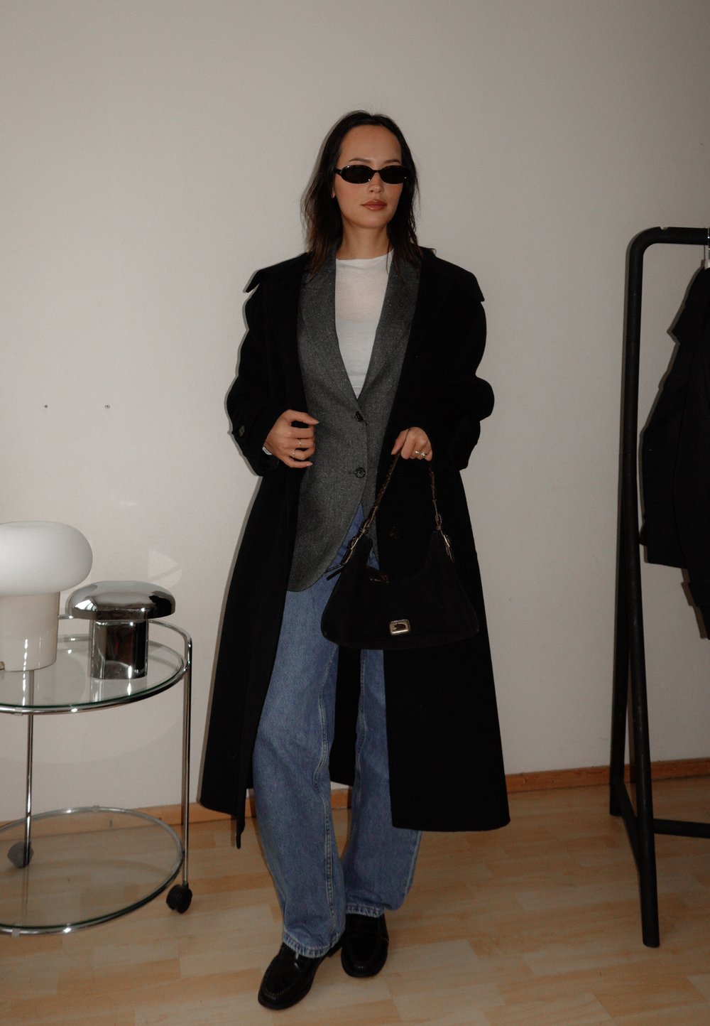 Dolg črn plašč čez sivo blazer, belo srajco in modre kavbojke. Dodatki vključujejo črne sončne očala in črno torbico. V bližini je krožna svetilka.