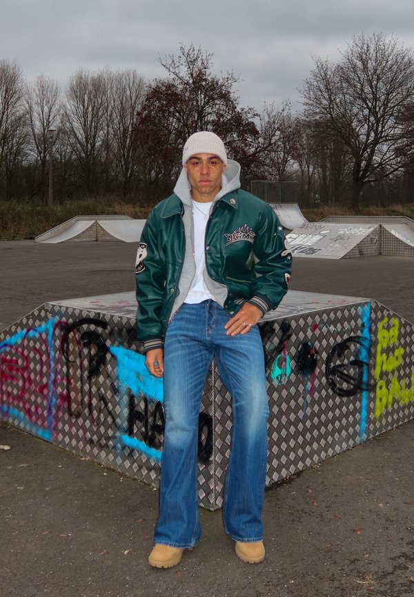 Zelená varsity bunda s šedou mikinou, bielym tričkom, rozšírenými modrými džínsami a béžovými topánkami, pózujúca pri kovovej skate rampě s graffiti.