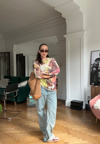 Mujer con gafas de sol, camisa de manga larga con flores, vaqueros holgados y sandalias, de pie en interiores con los brazos cruzados y llevando un gran bolso marrón en el hombro.