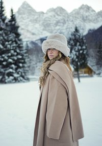 Femme portant un manteau beige et un chapeau en fourrure, debout dans un paysage enneigé avec des pins et des montagnes en arrière-plan.