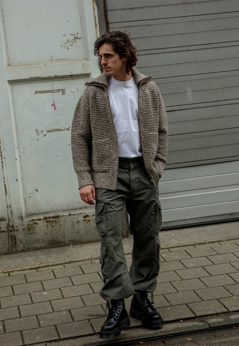 Storos mezginiai šviesiai rudas kardiganas su aukštu apykaklę, balta marškinėliai, alyvuogių žalios spalvos krovinių kelnės ir juodi bateliai su raišteliais ant akmenų grindinio.