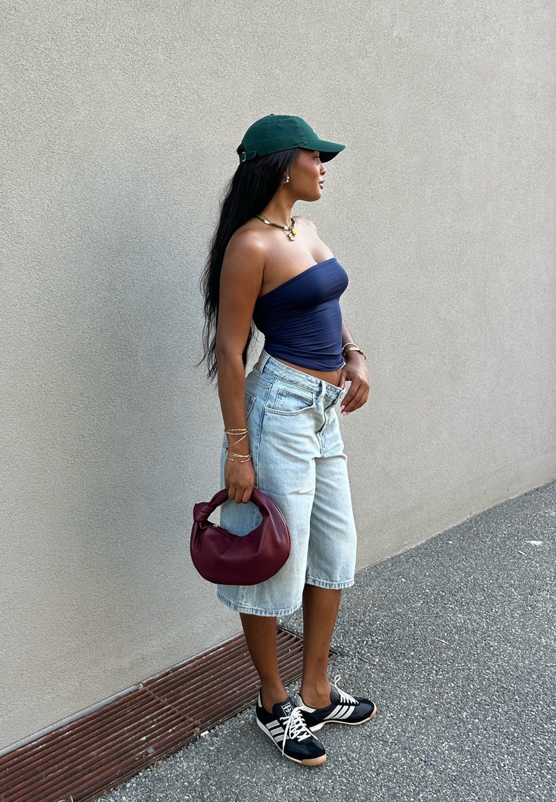 Top strapless azul marinho, calções Bermuda em denim azul claro, ténis preto e branco, segurando uma mala texturizada em bordeaux, adornada com pulseiras douradas.