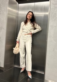 Balta megzta kardiganas su apvaliu iškirpimu, derinamas su baltais plačiakulkšniais pantalonais. Sidabriniai smailianosiai bateliai ir pūkuotas kreminis krepšys. Liftas kaip fonas.