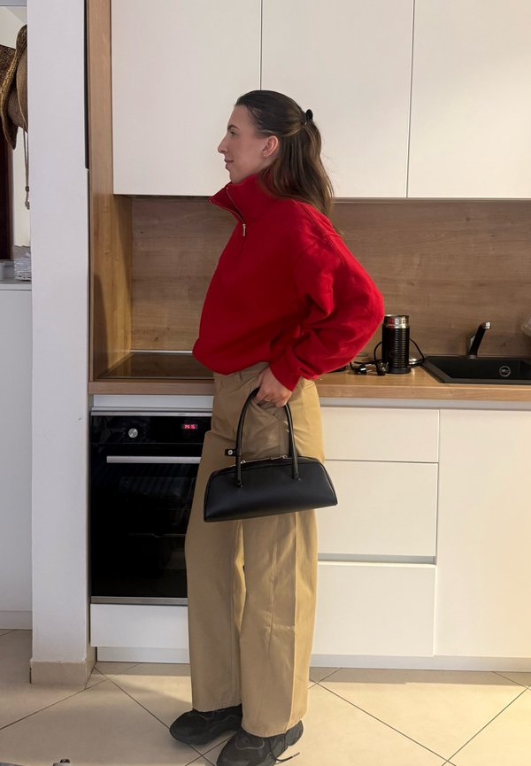 Una mujer con un jersey rojo y unos pantalones beige sostiene un bolso negro en una cocina moderna.