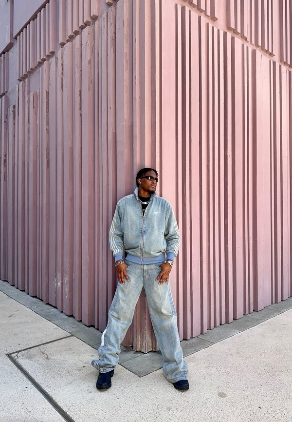 Homme portant un survêtement en denim et des lunettes de soleil appuyé contre un mur d'angle haut rose rainuré sur un trottoir en béton.