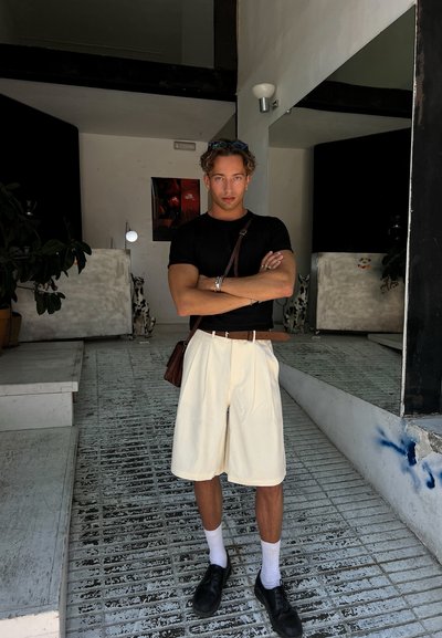 Pantalones cortos anchos de color claro, camiseta ajustada negra, cinturón marrón, zapatos negros y calcetines blancos. Ambiente interior con espejos y plantas.