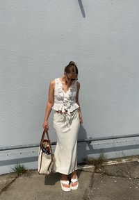 Weißes, gerafftes Crop-Top mit V-Ausschnitt, kombiniert mit einem langen weißen Rock mit Zugband. Das Model hält eine Tasche mit braunen Akzenten. Weiße Sandalen.