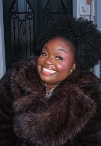 Χαμογελαστό άτομο με φυσικά σγουρά μαλλιά που φοράει σκούρο καφέ γούνινο παλτό και χρυσά κρίκους σκουλαρίκια, στέκεται μπροστά σε μια μαύρη πόρτα.