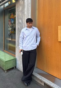 Camicia a righe blu chiaro con bottoni, pantaloni a gamba larga neri, che tiene una tazza di caffè, in piedi contro un muro di legno vicino a una finestra di vetro.