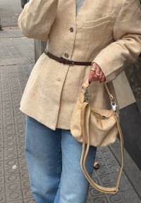 Personne portant un manteau beige tacheté avec une ceinture marron, un jean bleu, et tenant un sac à main beige sur un trottoir en ville.