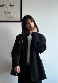 Veste noire surdimensionnée avec une finition texturée, portée sur un haut blanc, associée à une jupe à motif à pois. Sac noir avec bandoulière.
