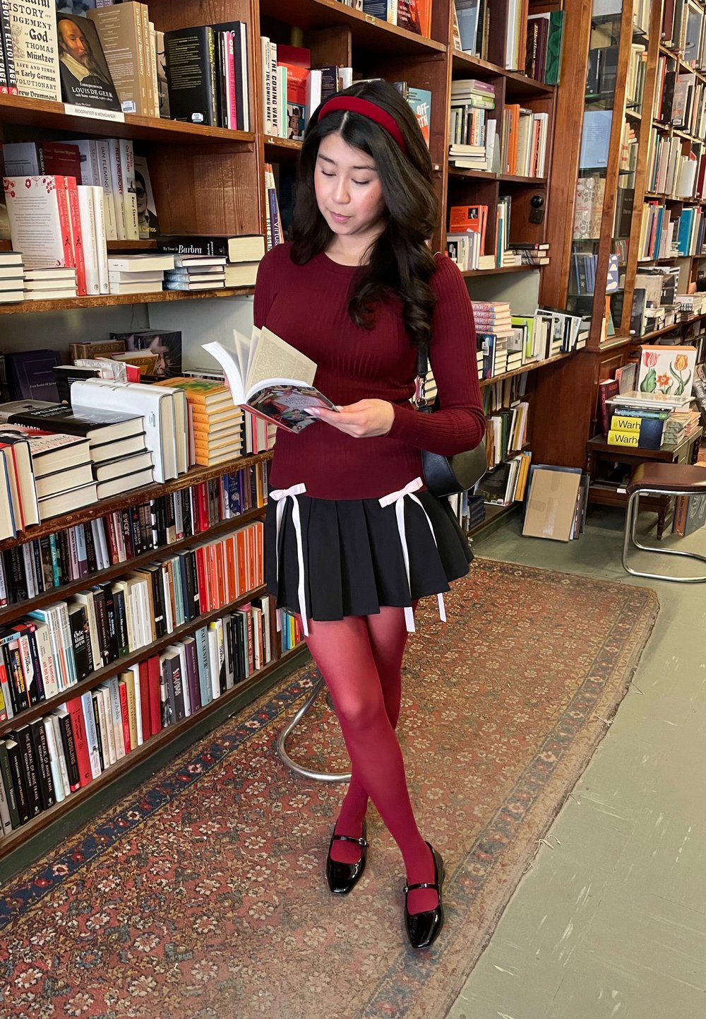 Blusa roja de manga larga, falda negra con detalles en cinta blanca, pantys rojos y zapatos negros. De pie en una biblioteca, sosteniendo un libro abierto.