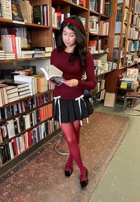 Blusa roja de manga larga, falda negra con detalles en cinta blanca, pantys rojos y zapatos negros. De pie en una biblioteca, sosteniendo un libro abierto.