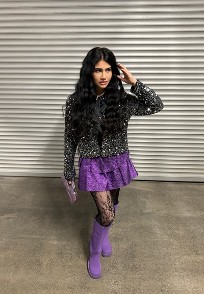 Chaqueta de lentejuelas negras brillantes, falda morada con volantes, medias de encaje negras y botas moradas hasta la rodilla; sosteniendo un pequeño bolso morado.