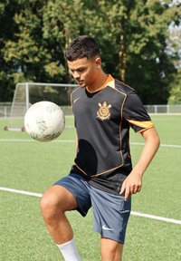 Černý dres s oranžovými detaily, logo na prsou, spárovaný se šedými šortkami a bílými ponožkami; hráč balancuje bílý fotbalový mí�č na svém koleni.