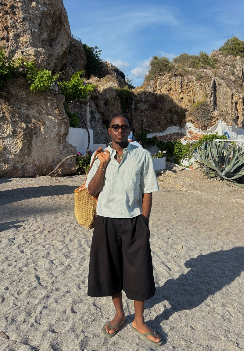 Hombre con gafas de sol, camisa ligera a rayas, pantalones cortos negros anchos y chanclas, sosteniendo una bolsa tejida en una playa de arena con acantilados rocosos detrás.