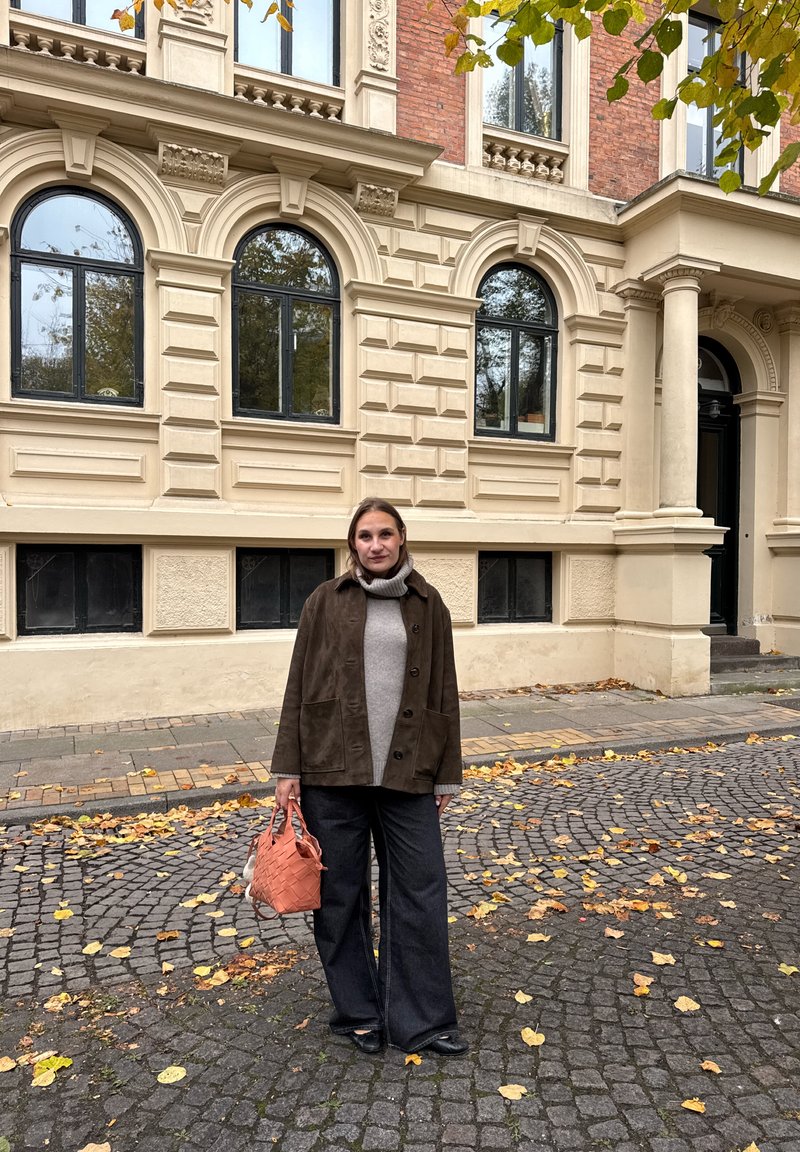 Bruine suède jas, grijze coltrui, zwarte wijde broek, en een oranje geweven handtas. Kasseienstraat met gevallen bladeren.