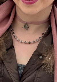 Colliers ton argent : l'un avec un pendentif en forme d'étoile et l'autre avec des anneaux métalliques reliés. Le modèle porte un collier ras du cou rose et une veste marron avec des accents en fourrure.