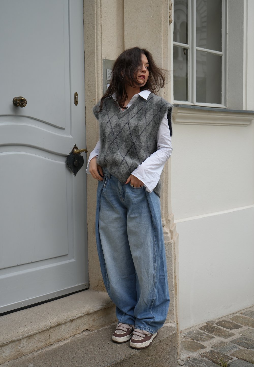Grijze en zwarte argyle vest over een wit shirt, gecombineerd met oversized lichtblauwe jeans en bruine sneakers, staande bij een grijze deur.