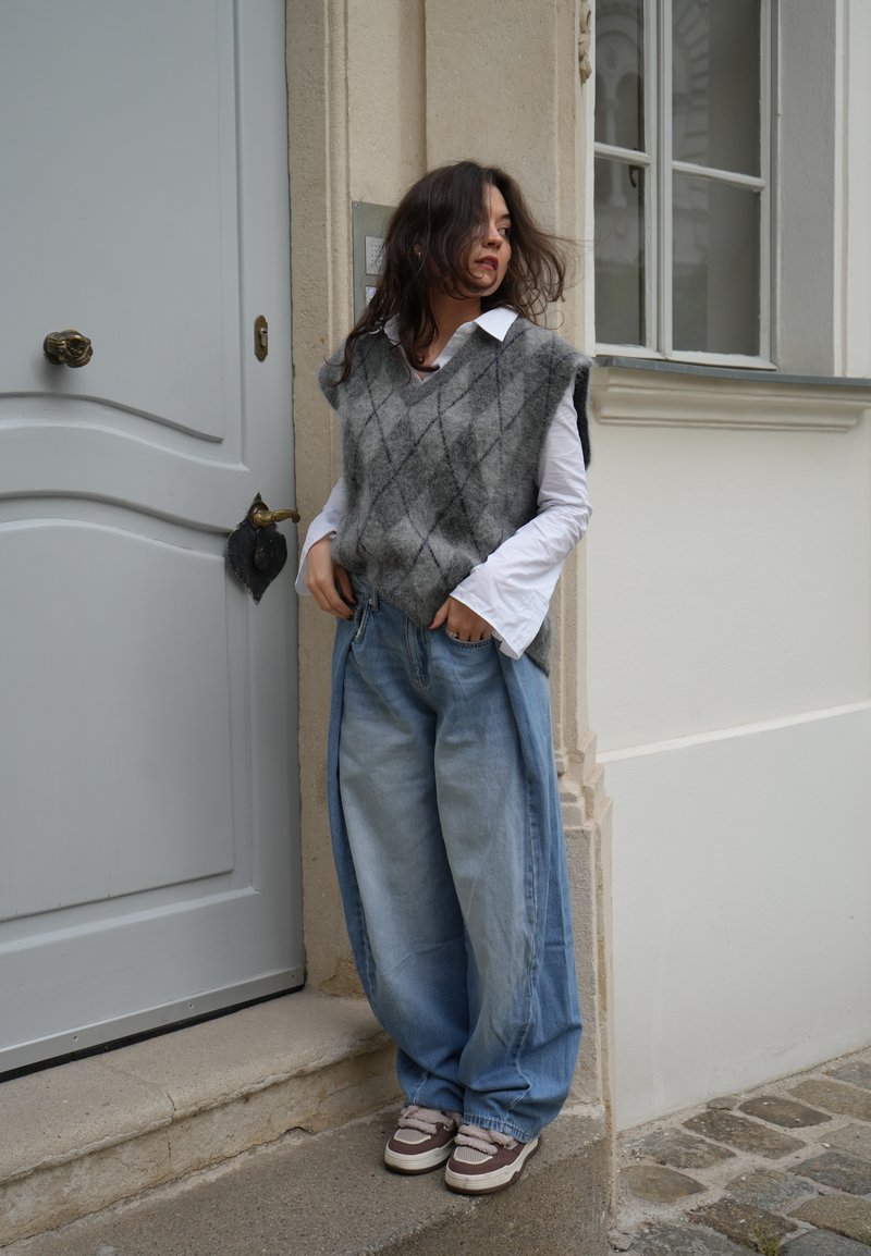 Šedý a černý argyle svetr přes bílou košili, spárovaný s oversized světle modrými džíny a hnědými teniskami, stojící u šedých dveří.