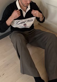 Chemise style rugby en noir et blanc avec le logo "Gant 1949", associée à un pantalon gris ample. Assis sur un sol en bois clair.