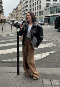 Fekete bőrkabát cipzáras záródással, fehér ing és barna, bő szárú nadrág. Fekete napszemüveget tart a kezében, valamint van egy bézs színű, szövött táskája.