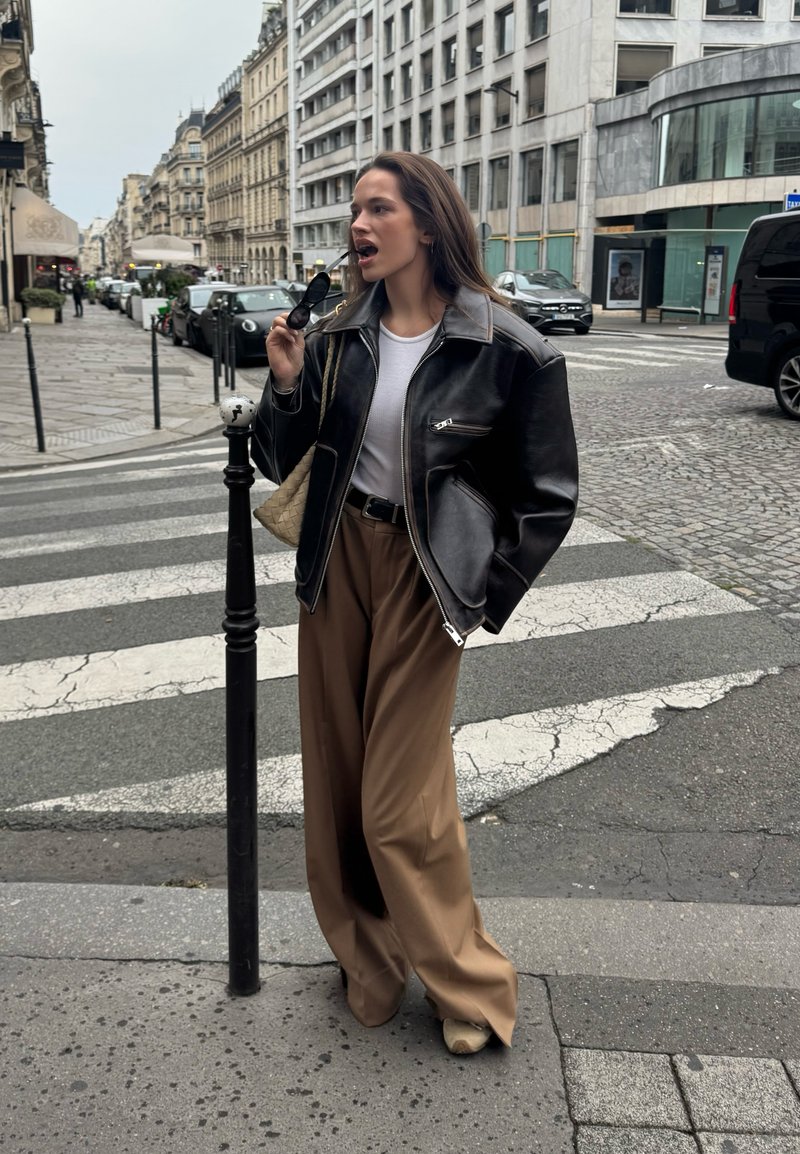 Black leather jacket with a zip closure, white shirt, and brown wide-leg trousers. Holding black sunglasses, with a woven beige bag.