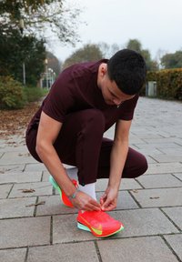Chaussures de course Nike orange néon avec des accents verts et un matériau en maille léger. Une personne fait les lacets en portant une tenue de sport bordeaux.