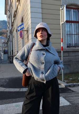 Mujer con cárdigan gris de punto y sombrero, pantalones negros de rayas, bolso marrón colgado al hombro, de pie en una calle de la ciudad cerca de señales de tráfico.