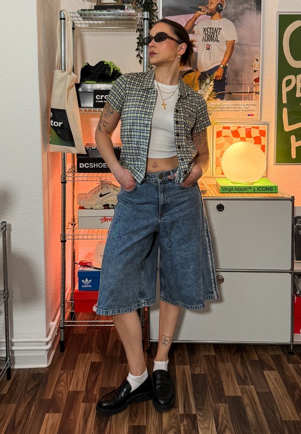 Persoon met een zonnebril, een kortemouwen geruit overhemd over een wit crop top, losse denimshorts en zwarte schoenen, staand in een versierde kamer.