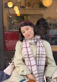 Beige Trenchoat, pinkes und braunes kariertes Tuch, rosa Handtasche. Weiche Texturen, lässiges Design. Holz-Hintergrund mit warmen hängenden Lichtern.