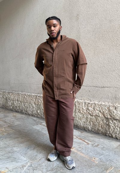 Camisa de botón marrón con un corte relajado y mangas cortas, combinada con pantalones marrones a juego y zapatillas grises, frente a una pared texturizada.