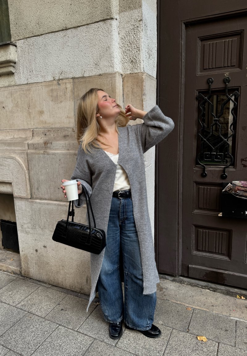 Hellgrauer, langer Cardigan über einem weißen Oberteil, kombiniert mit weiten blauen Jeans. Hält eine weiße Tasse und eine schwarze, strukturierte Handtasche. Schwarze Schuhe.