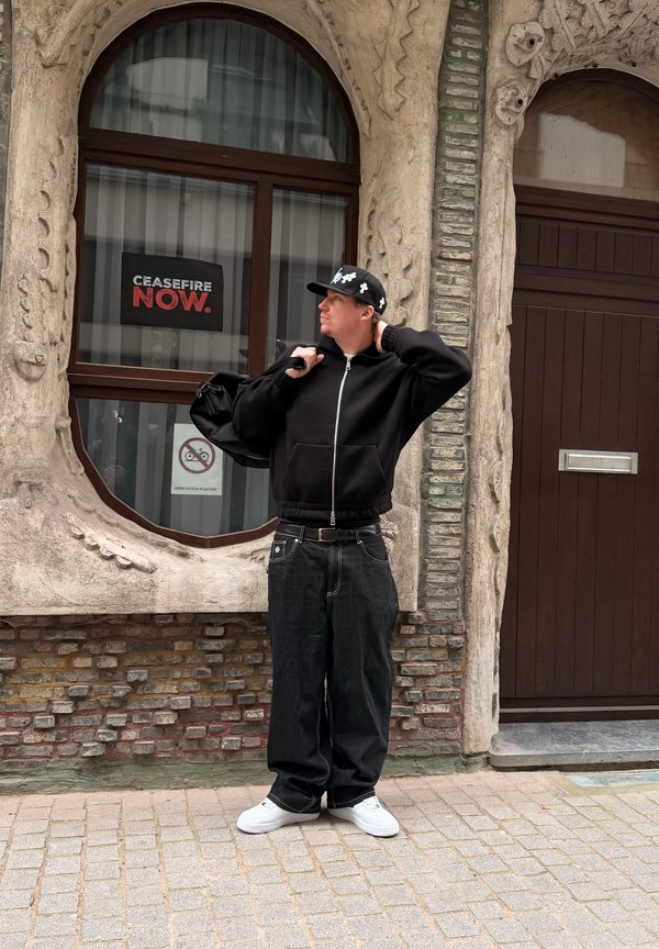 Hombre con sudadera negra con capucha, gorra negra y mochila al hombro, de pie en una calle empedrada junto a una pared de piedra ornamentada con una ventana que muestra un cartel con el texto "ALTO AL FUEGO AHORA".