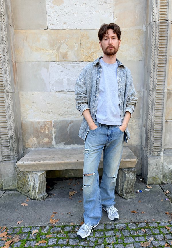 Hombre con barba y cabello oscuro, de pie con las manos en los bolsillos cerca de un banco de piedra y una pared, vistiendo una camisa gris, chaqueta clara, vaqueros rotos y zapatillas deportivas.